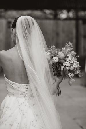 A bride in a white wedding dress with delicate lace details and flowing veil holds a romantic floral bouquet.
