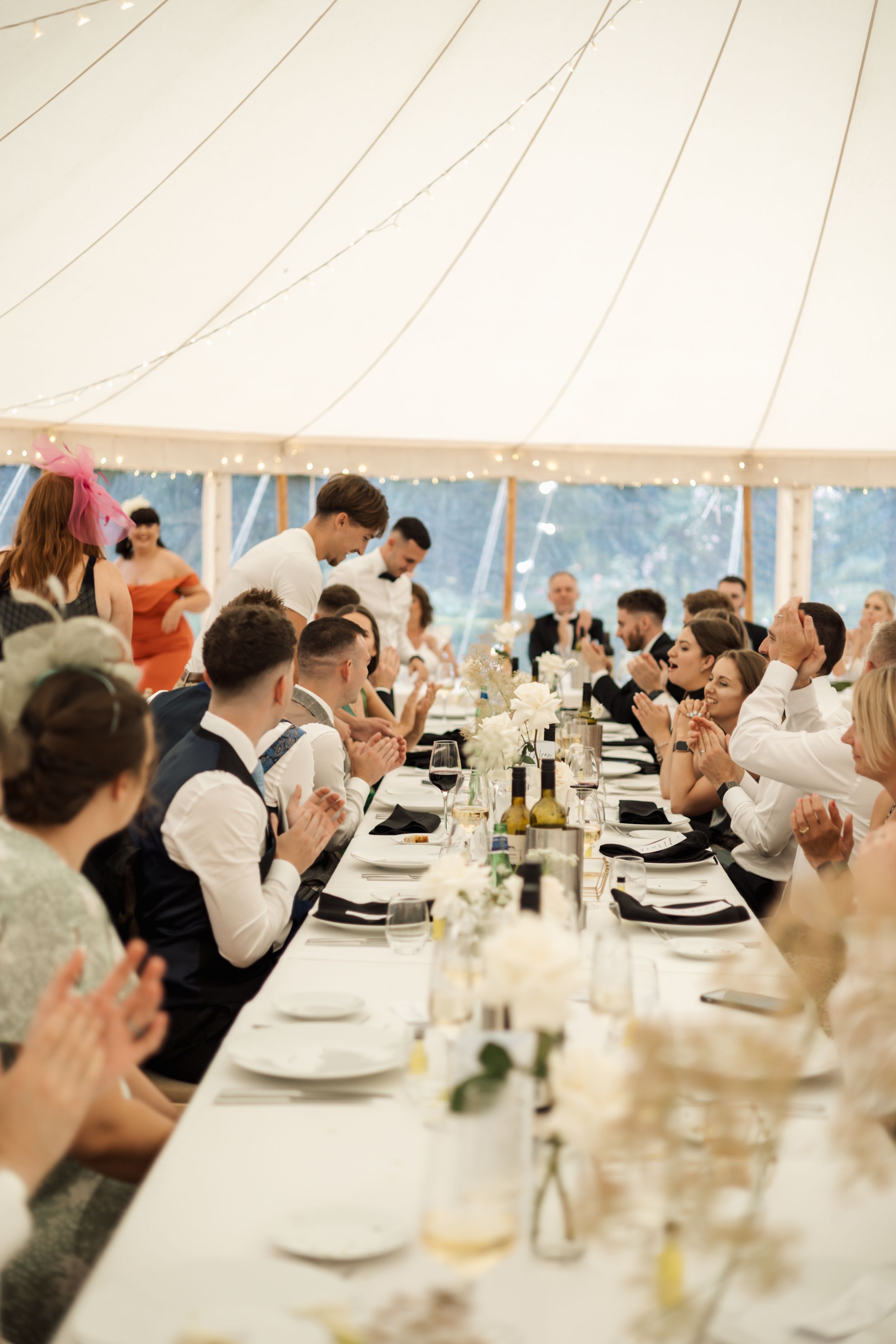 Wedding reception dinner under a white tent with long tables and festive lighting.