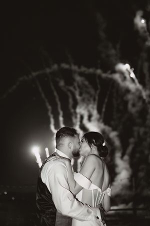 A romantic nighttime wedding kiss is captured under string lights with fireworks in the background.