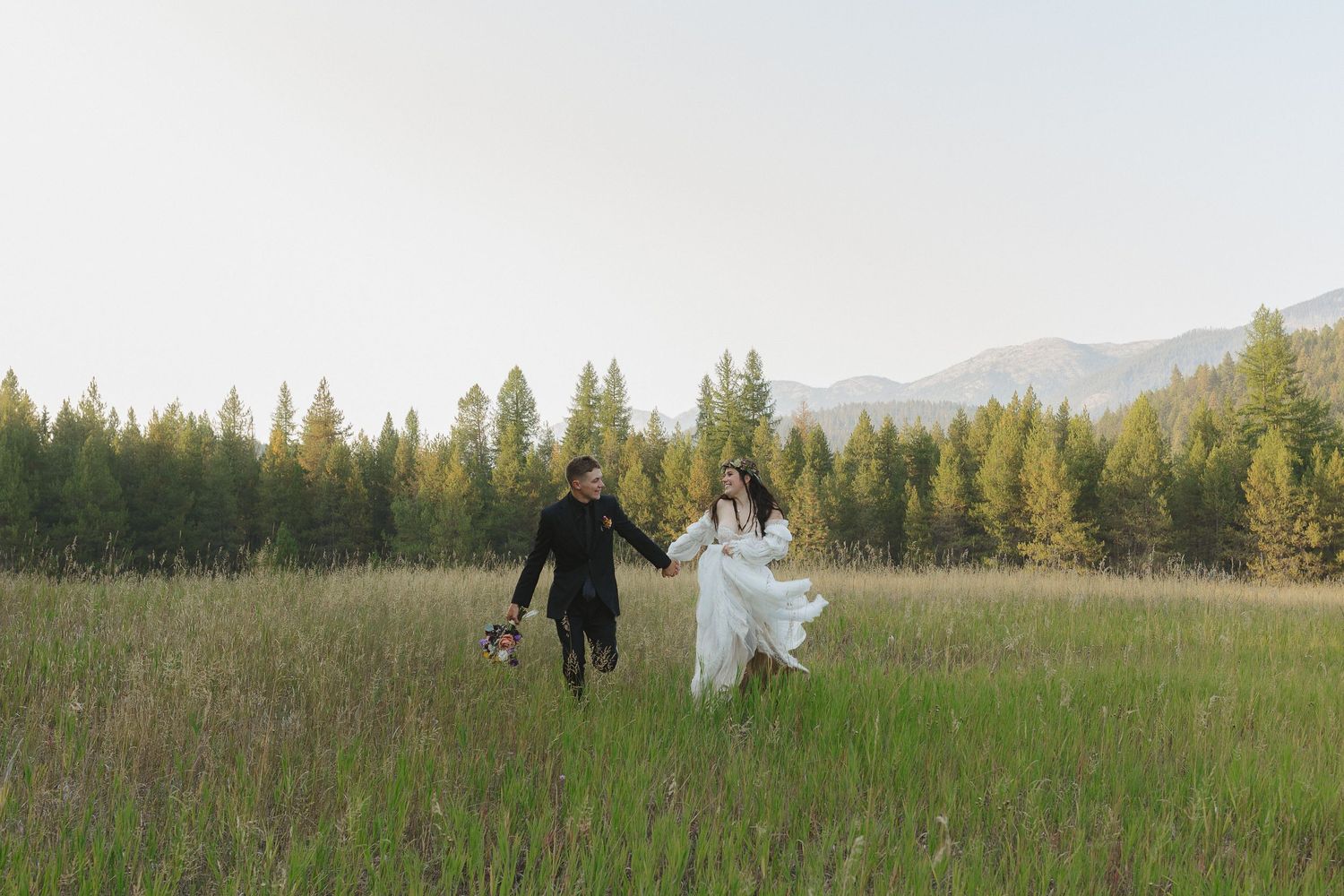 Couple in wedding attire runs joyfully through a grassy meadow with evergreen trees and mountains in the background.
