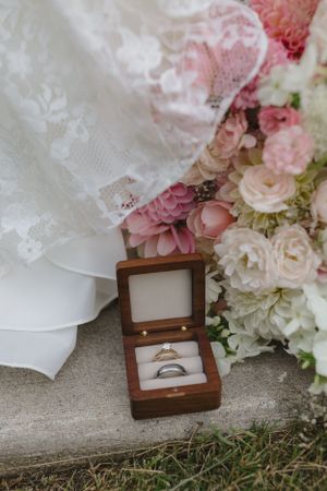 Wedding rings in a wooden box next to delicate lace fabric and pink and white floral bouquet on stone surface.