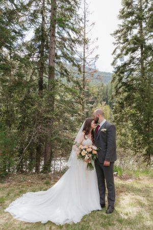 A romantic wedding couple stands among tall evergreen trees in a mountainous forest setting with flowing white gown.