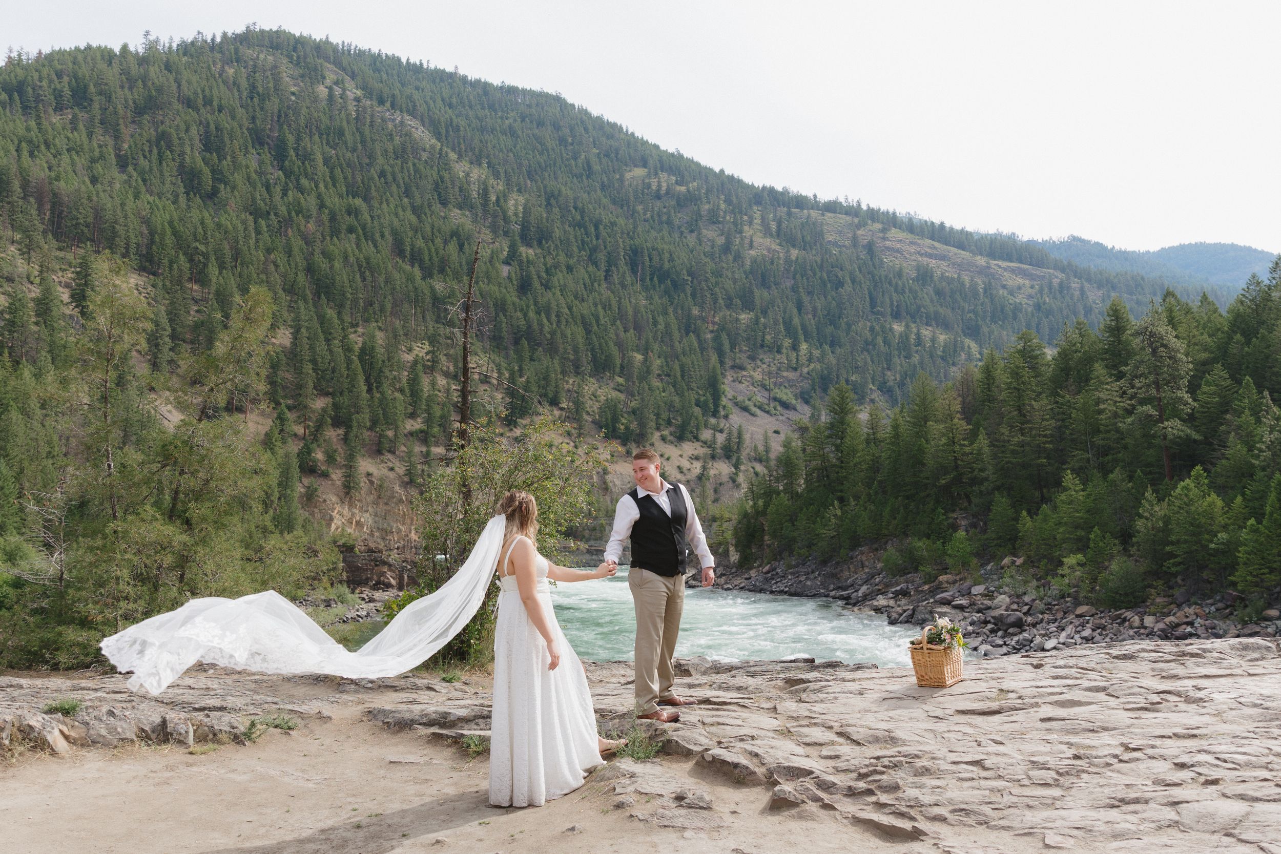 A couple in wedding attire stands beside a scenic mountain river as a flowing veil catches the breeze.
