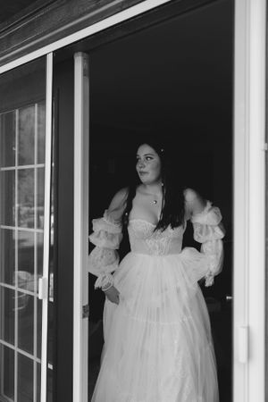 A black and white photograph shows someone in an elegant white vintage-style dress with puffy sleeves standing in a doorway.