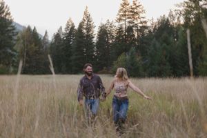 Two people walking together through a golden meadow surrounded by tall evergreen trees at dusk.