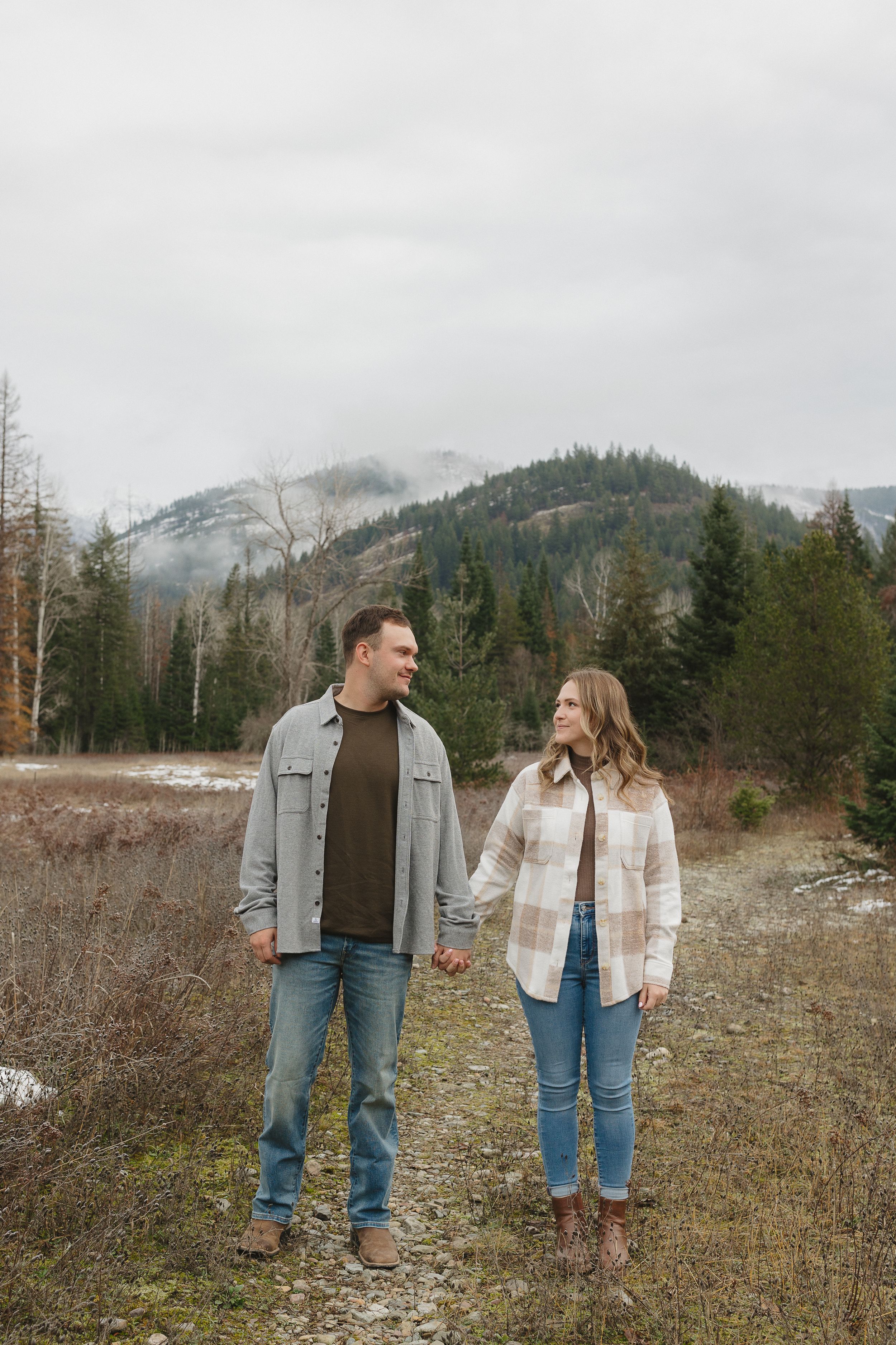A couple stands together in a meadow with snow-capped mountains and evergreen forest in the background on an overcast day.