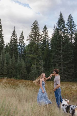 Couple dancing together in a grassy meadow surrounded by tall evergreen trees while their dog watches nearby.