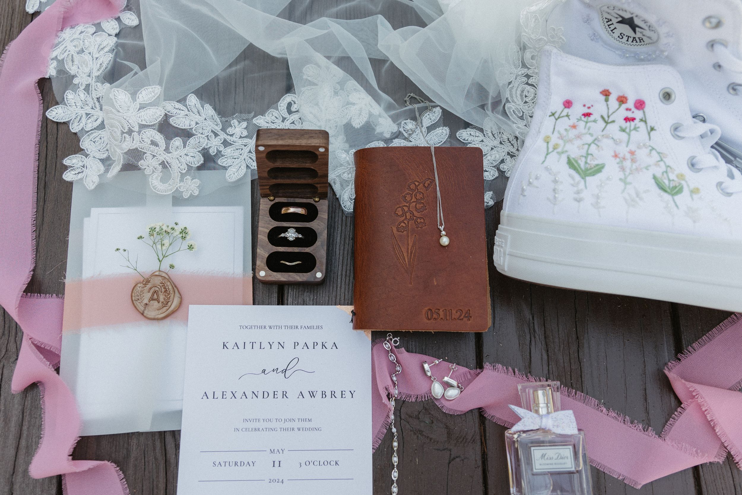 Wedding details flatlay with invitation, vintage camera, delicate lace veil, and pink silk ribbon on wooden surface.