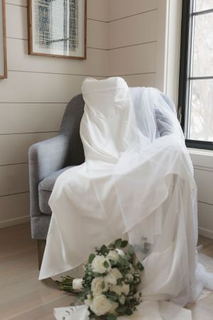 A white wedding dress draped over a gray armchair with a bridal bouquet of white and green flowers near a window.