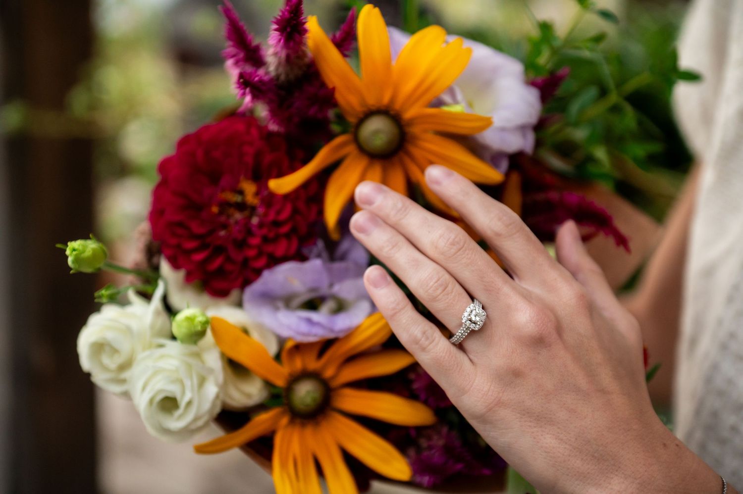 Hand wearing engagement ring holds vibrant bouquet with orange daisies, burgundy dahlias and white roses.
