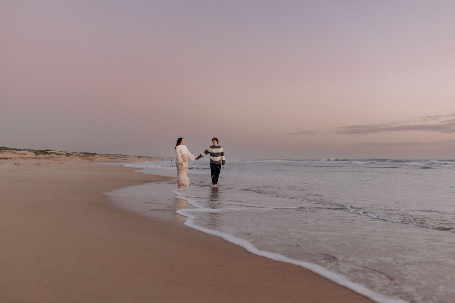 A pregnant person in white swimwear stands at the water's edge on a beach during sunset with soft pink clouds.