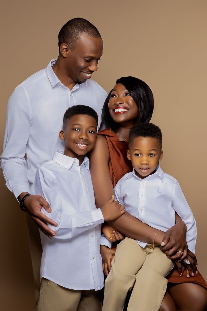 A loving family of four wearing coordinated white and tan outfits poses together against a brown backdrop.
