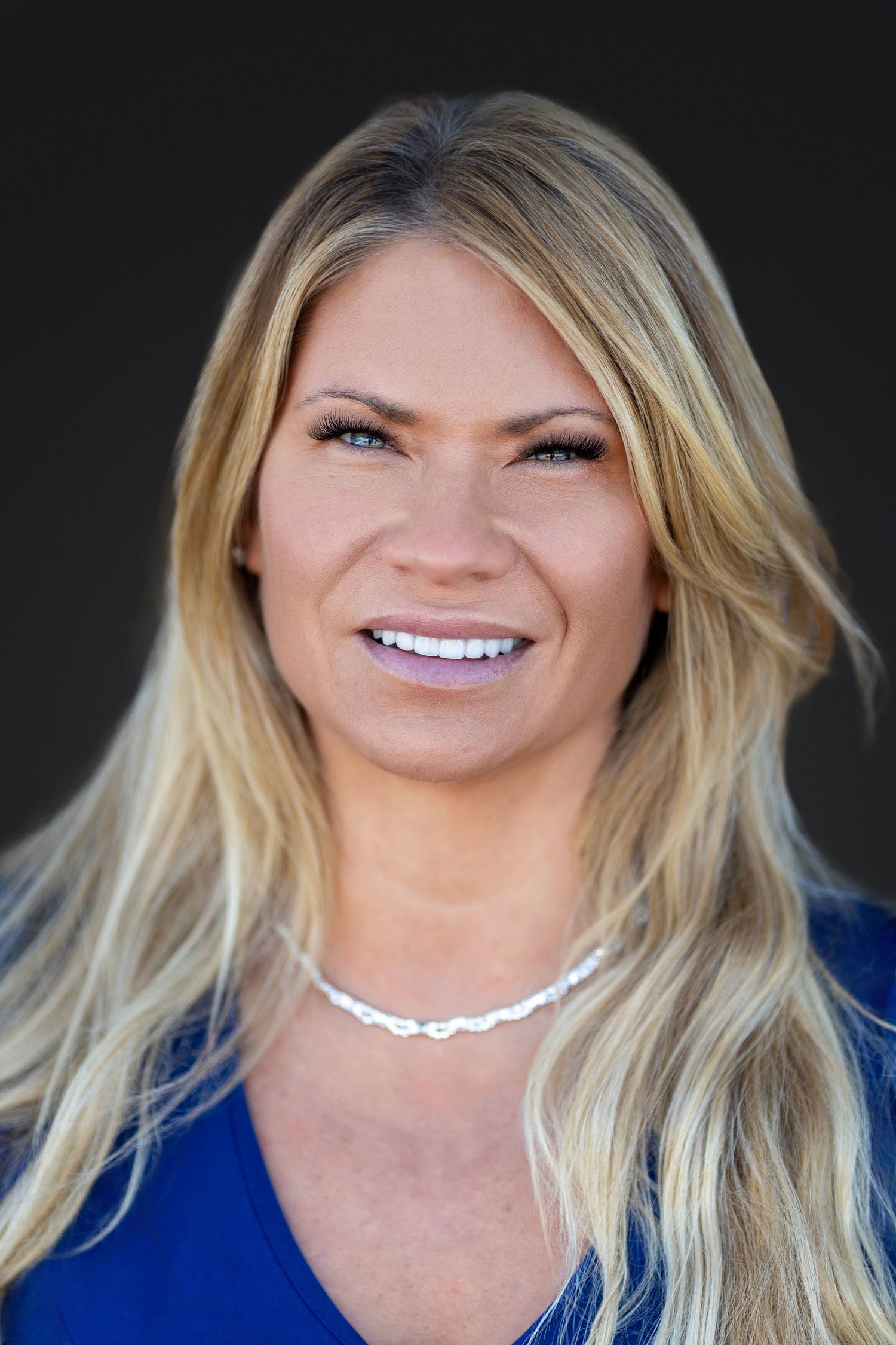 Professional headshot of someone with long blonde hair wearing a bright blue top and delicate chain necklace against dark background.