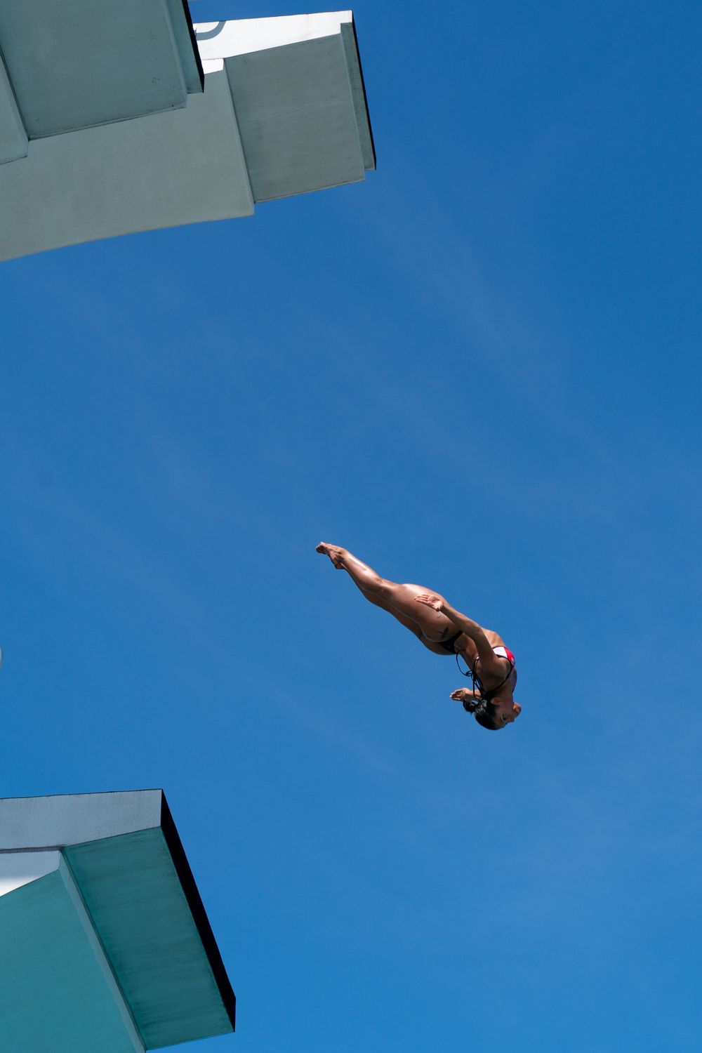 High diver in midair against a clear blue sky, captured as they leap from a diving platform during a sports and leisure photography