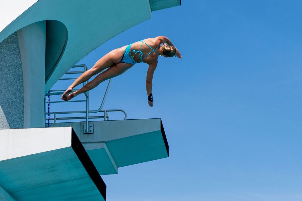 High diver in midair against a clear blue sky, captured as they leap from a diving platform during a sports and leisure photography
