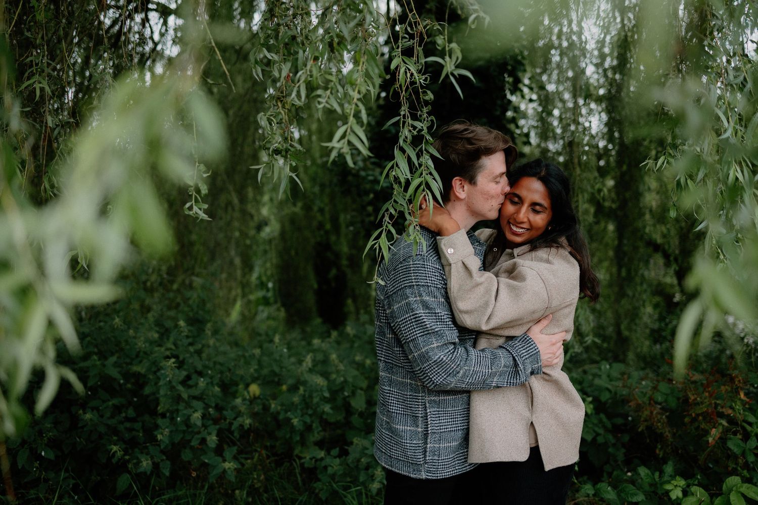 A couple shares an intimate moment while embracing under drooping willow tree branches.
