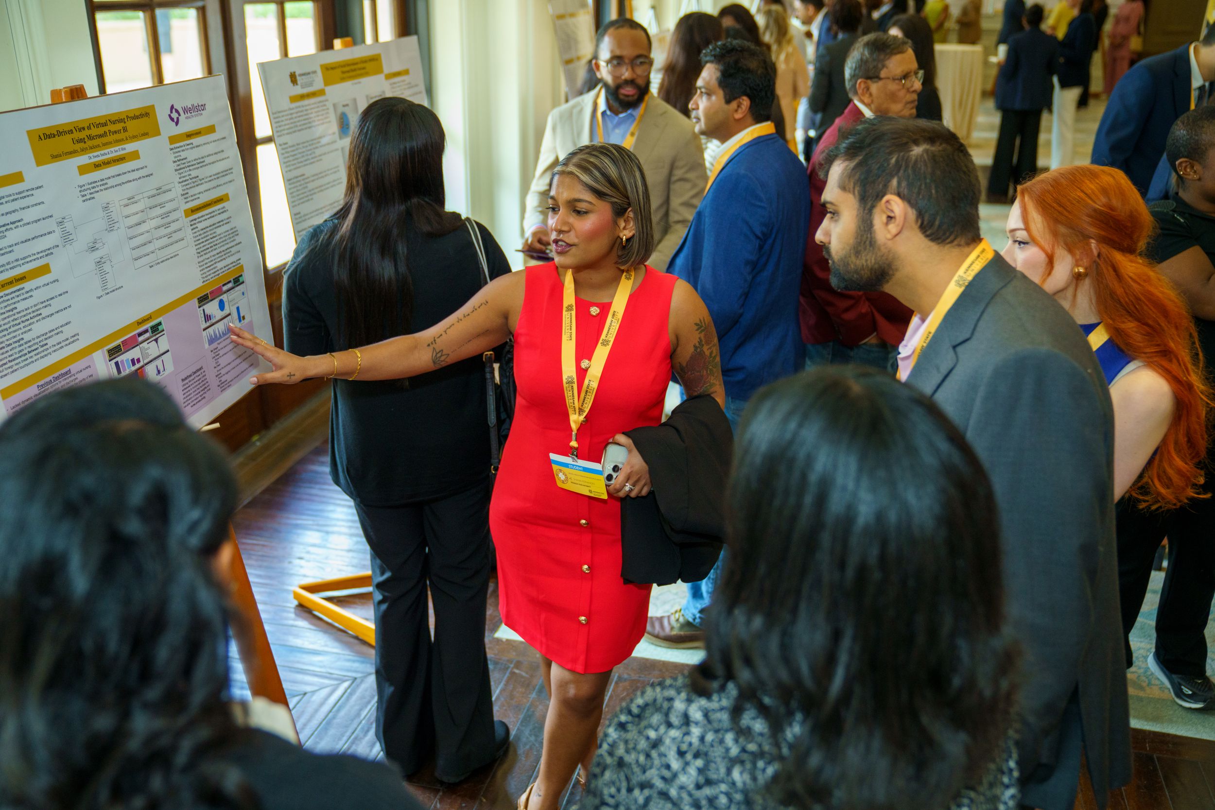A presenter in a red dress discusses a poster presentation at an academic conference with attendees in a busy hallway.