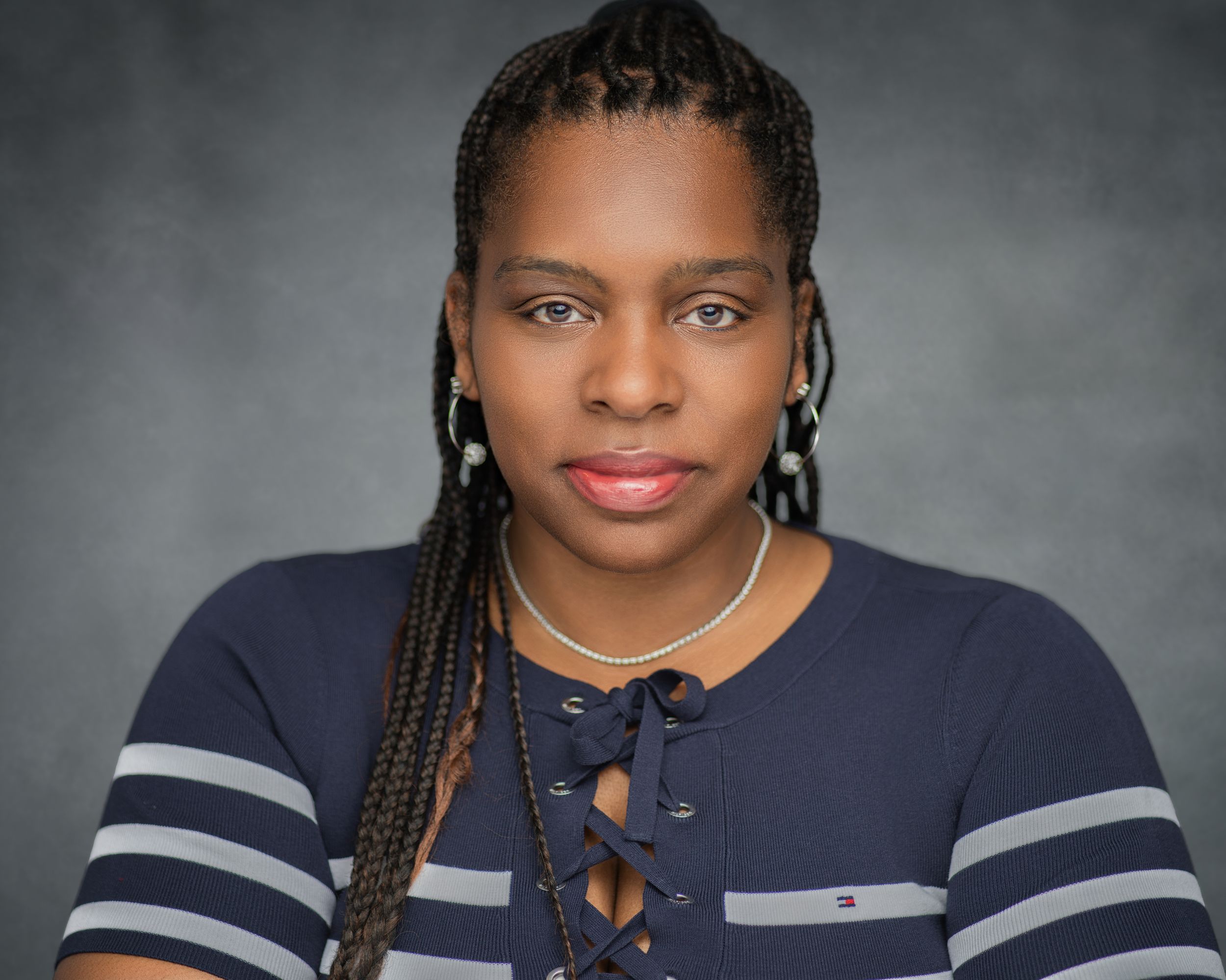 Professional headshot with braided hairstyle wearing a navy blue criss-cross tie sweater with white stripes and pearl earrings.