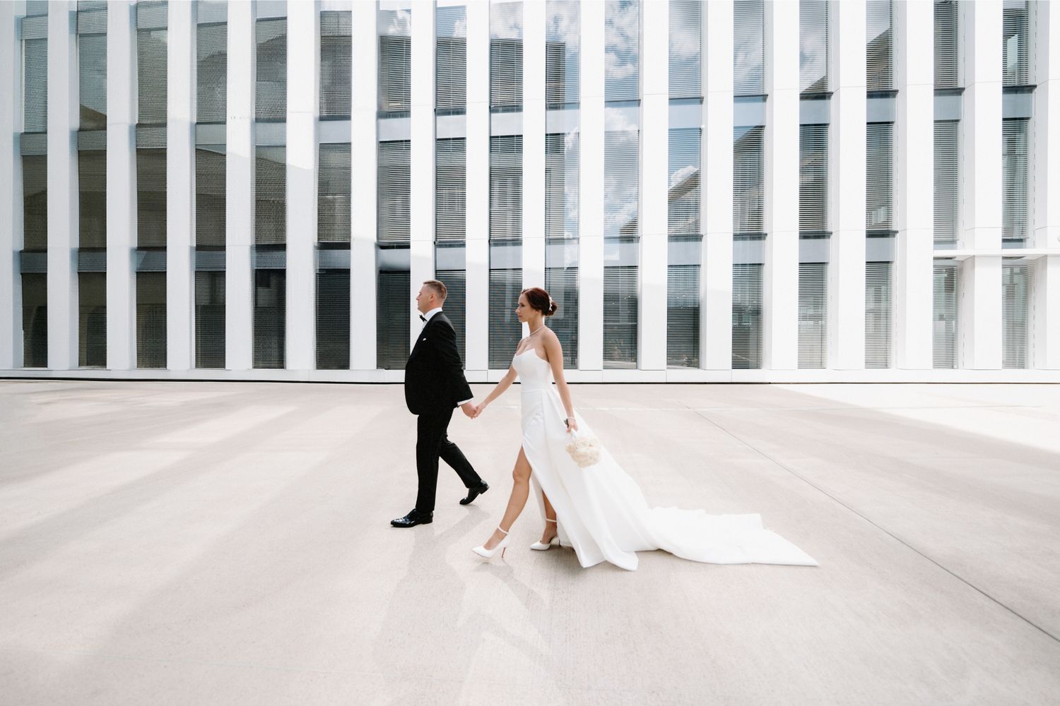 Couple walks hand in hand against modern architectural backdrop with flowing white dress.