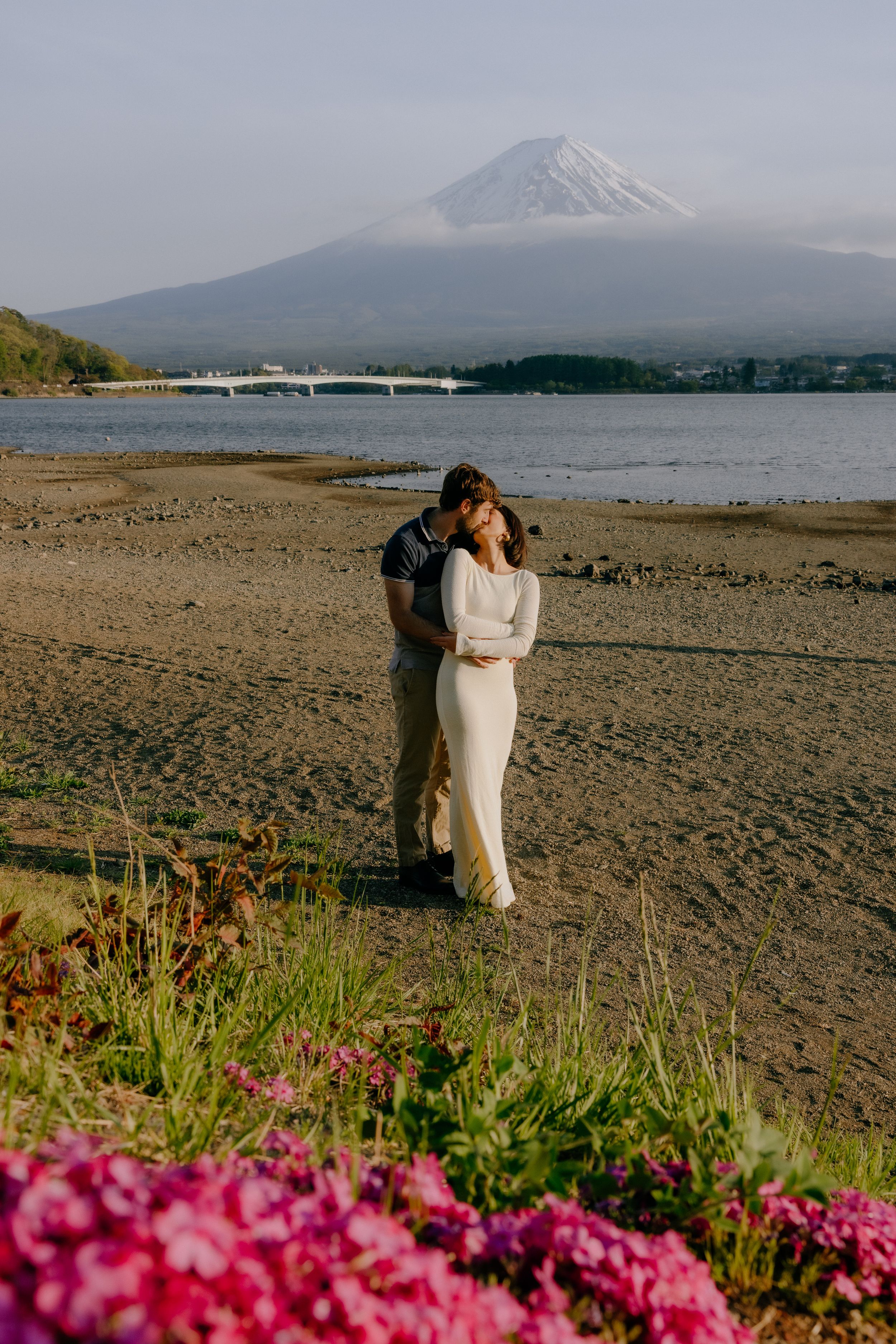 Scenic view of Mount Fuji with pink flowers in foreground and a couple embracing on a sandy beach by Lake Kawaguchiko.