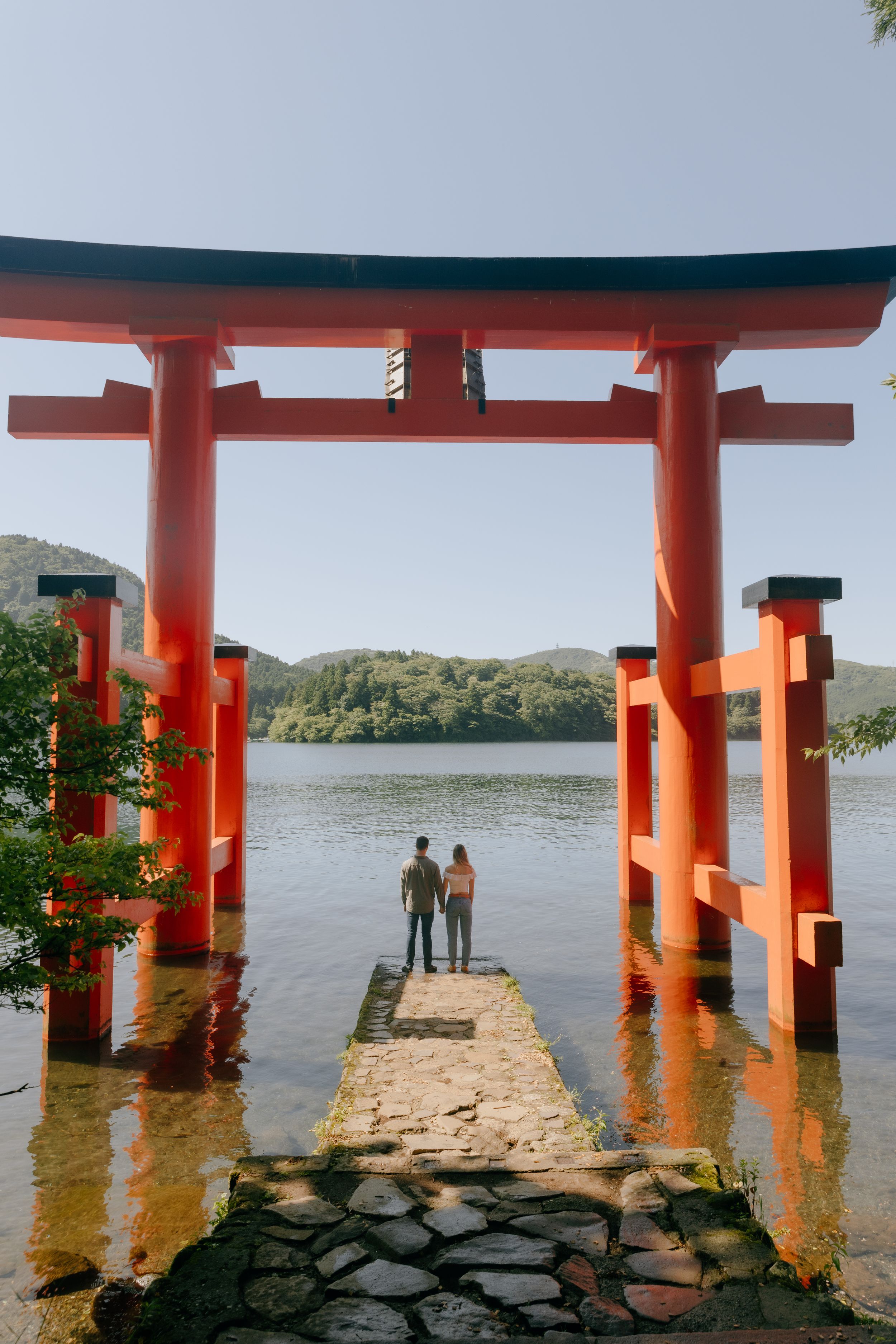 Traditional red Heiwa no torii gate with stone path leads into a tranquil lake surrounded by green hills in Hakone Japan.