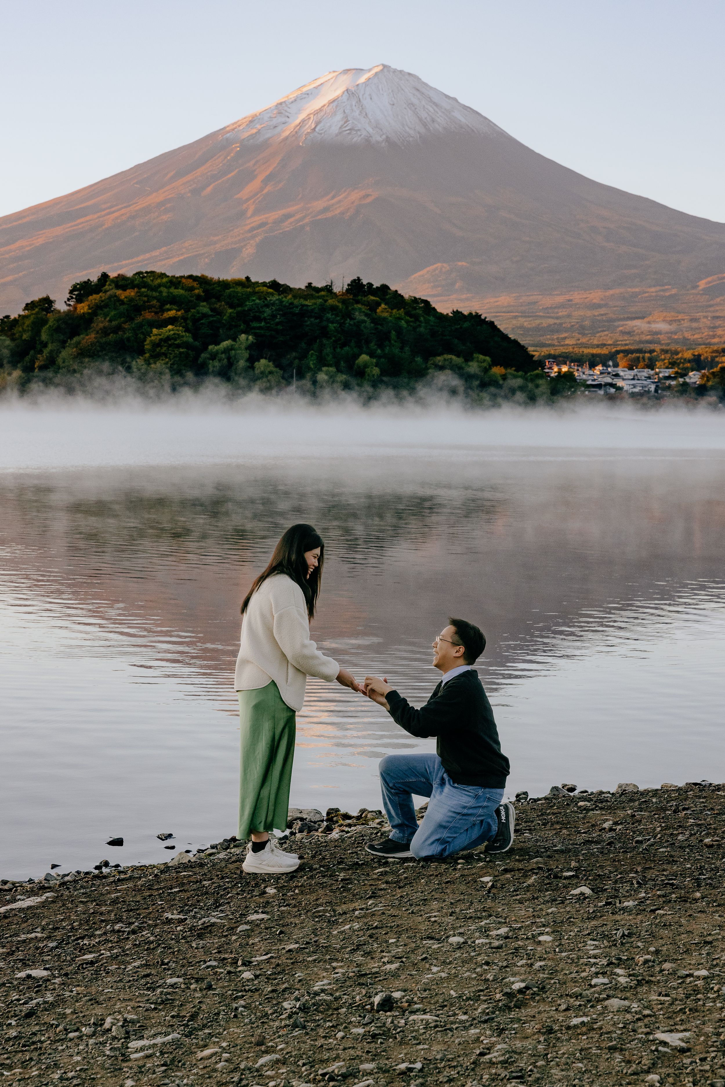 Two people sit by a misty lake with a mountain backdrop, creating a serene and romantic atmosphere in nature.