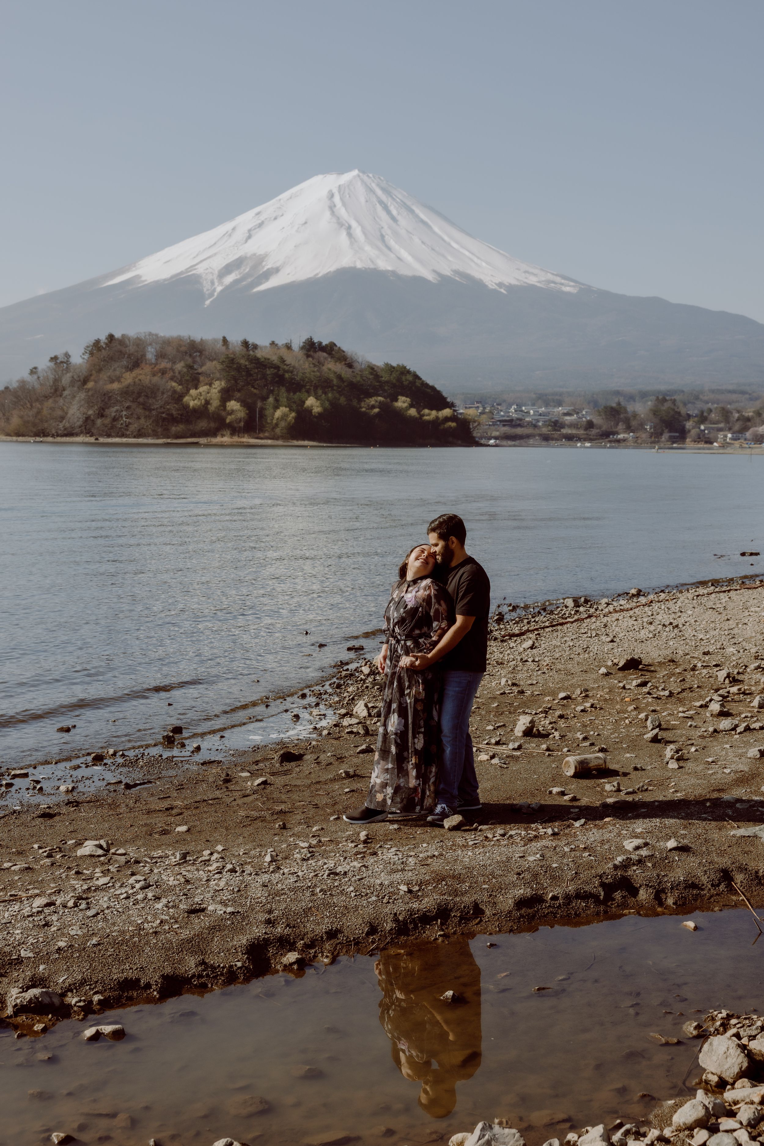 A couple stands on the rocky shores of Lake Kawaguchiko with majestic snow-capped Mount Fuji rising in the background.