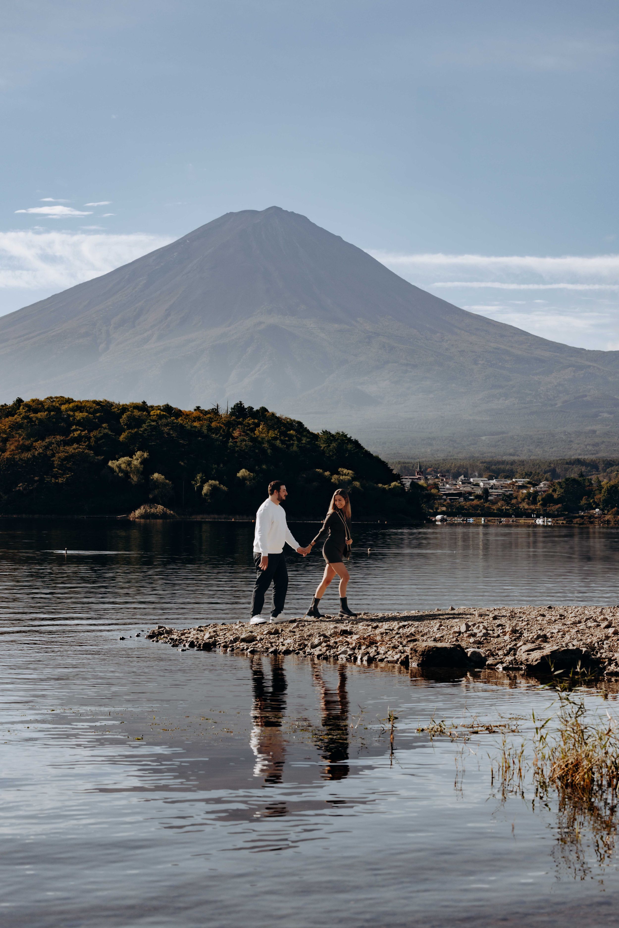 Two figures stand on a rocky peninsula with a dramatic mountain peak rising in the distance.