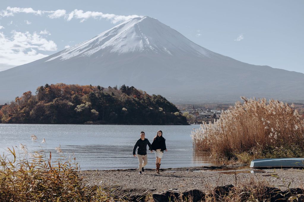 Two figures walk along the shore of Lake Kawaguchi with Mount Fuji's snow-capped peak rising majestically in the background.