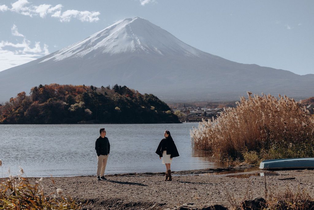 Two people stand by Lake Kawaguchiko with Mount Fuji rising majestically in the background on a clear autumn day.
