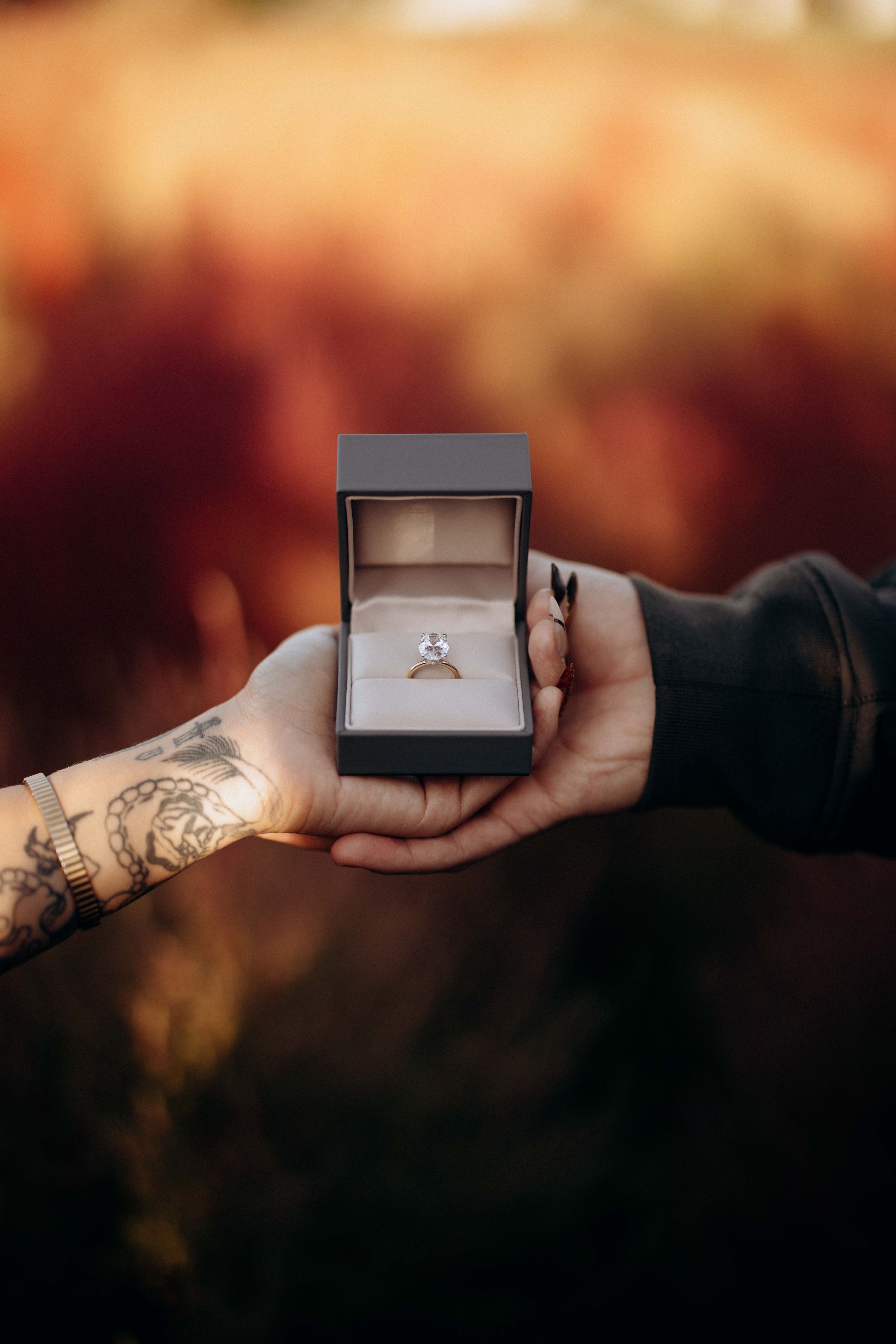 Tattooed hand holding a gray ring box with engagement ring against blurred autumn background.