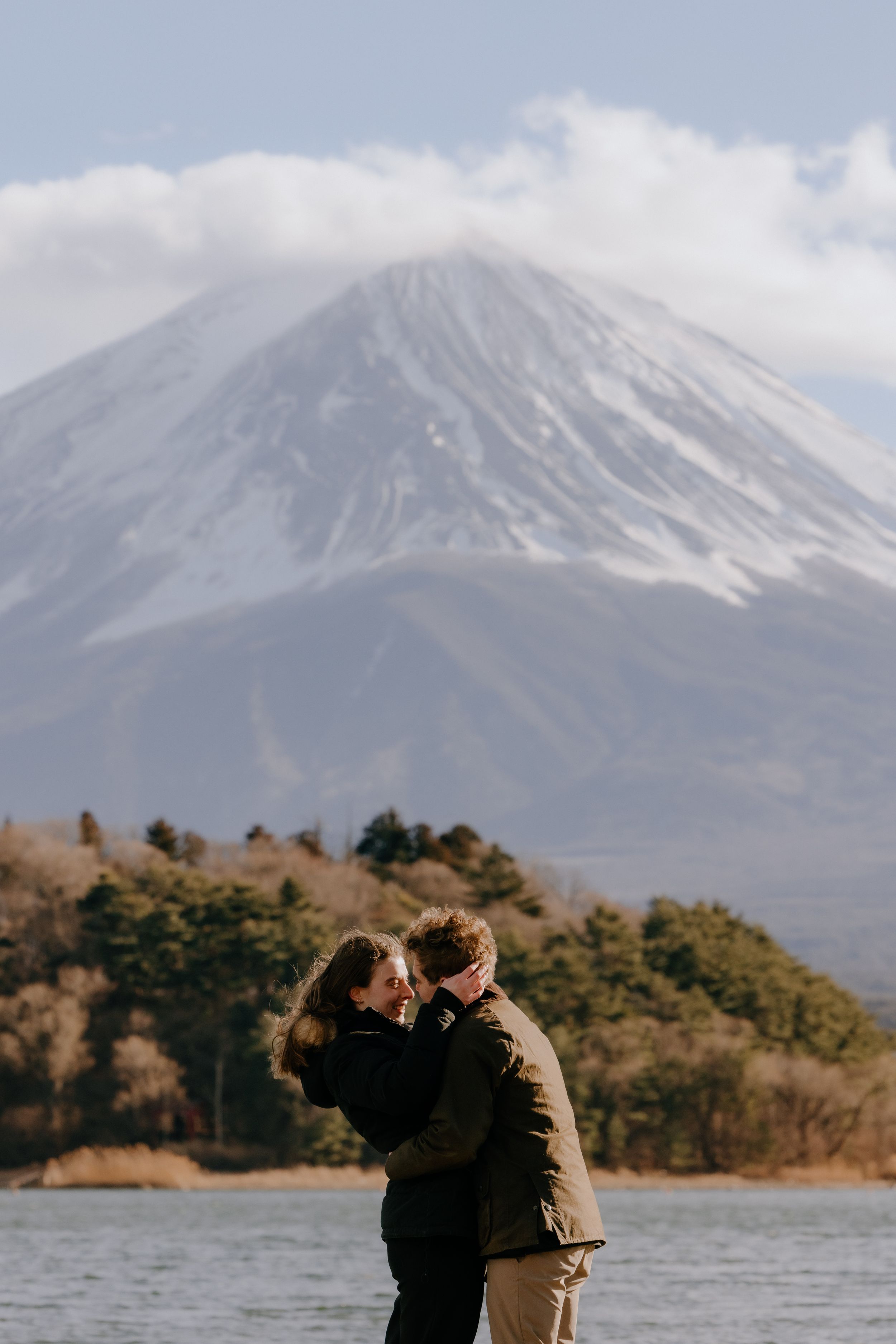 A couple embraces at Lake Kawaguchiko with snow-capped Mount Fuji rising majestically in the background on a cloudy day.