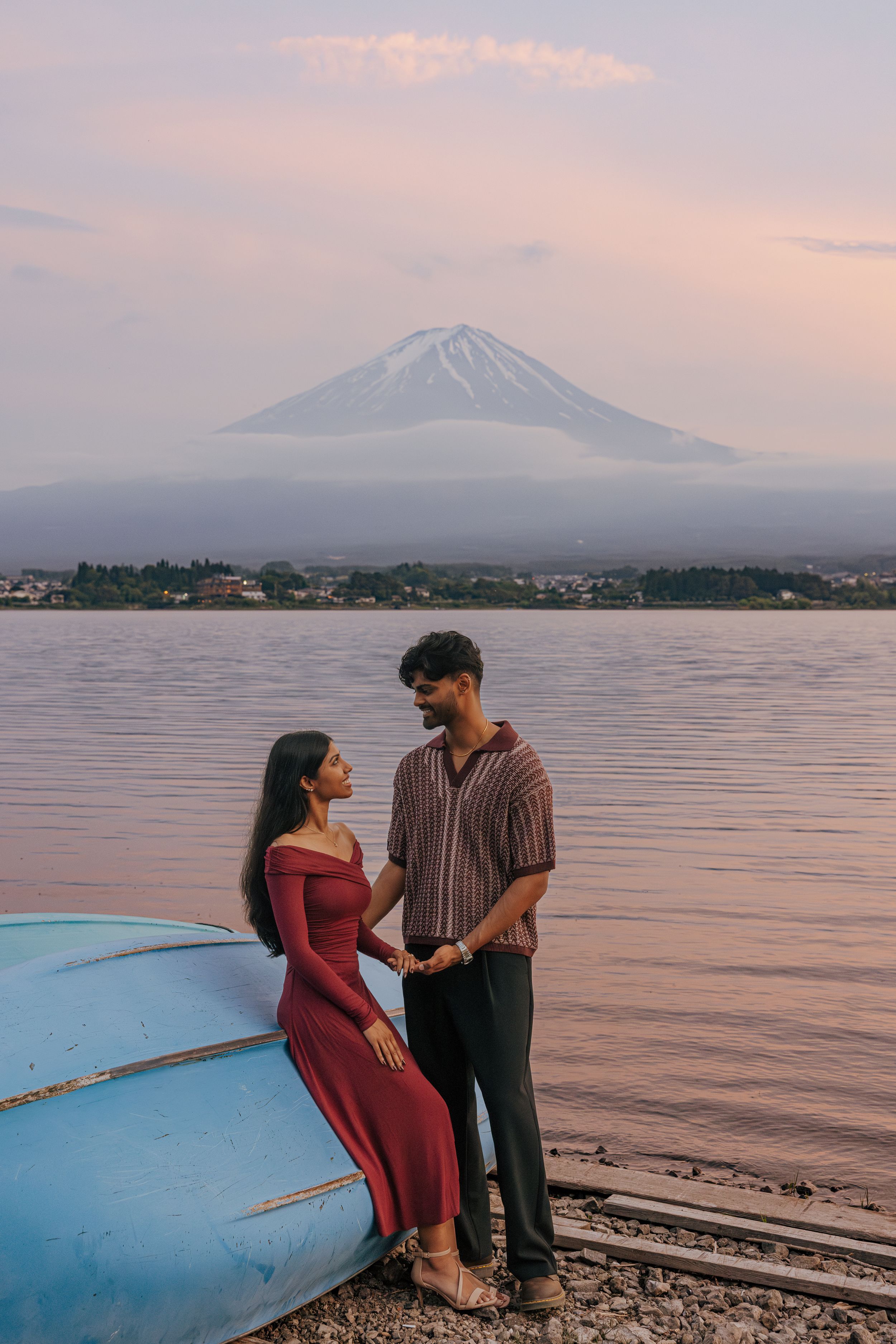 A couple stands by a blue boat at Lake Kawaguchiko with Mount Fuji visible in the misty sunset background.