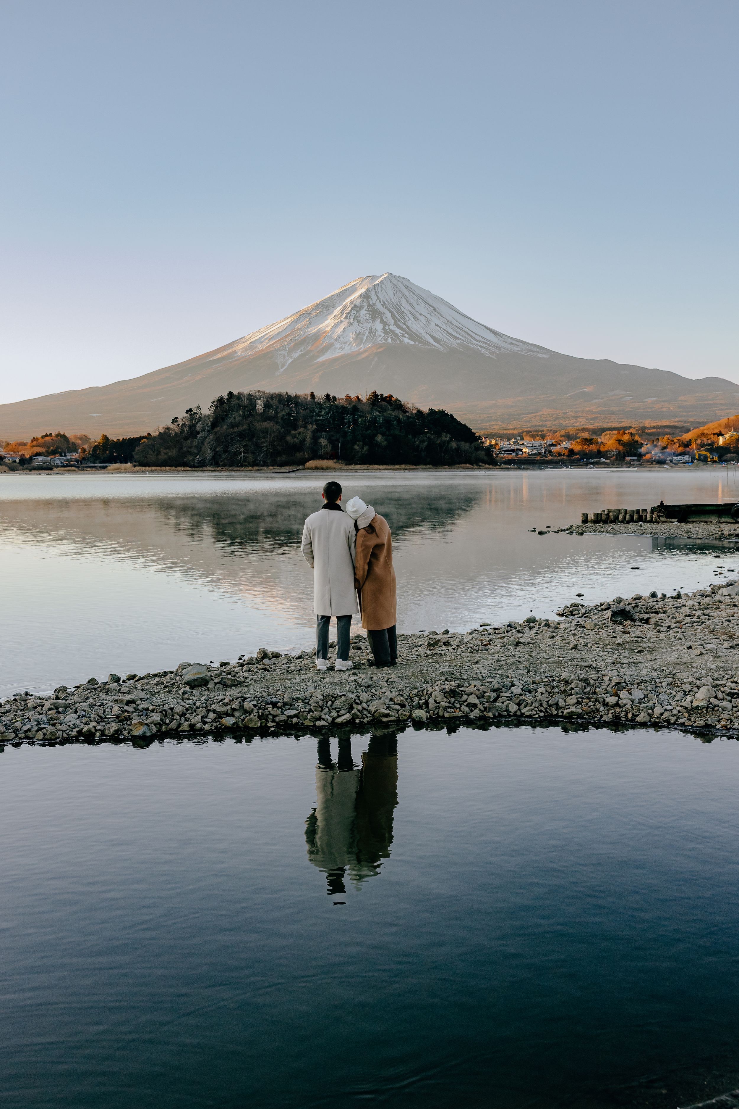 Mount Fuji reflects majestically in the calm waters of Lake Kawaguchiko at dawn, with figures silhouetted on the rocky shore.