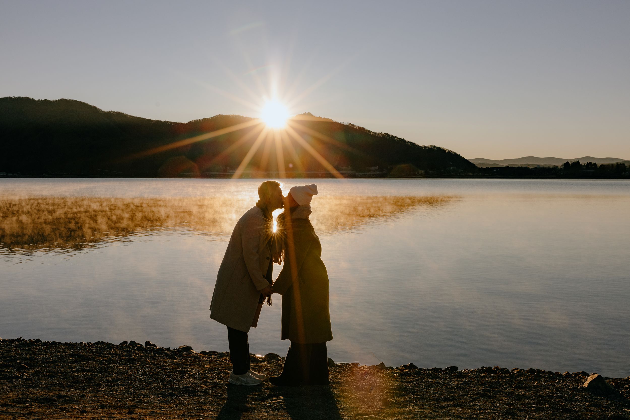 Silhouettes of a couple embrace against a dramatic sunset over a tranquil lake creating a romantic golden hour scene.