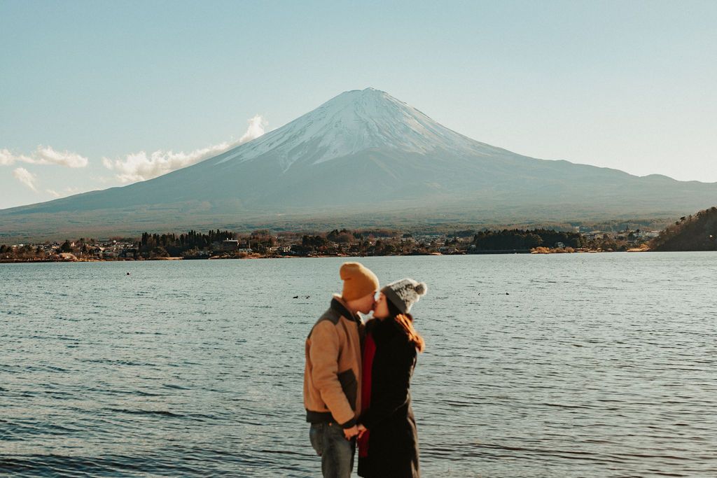 Two figures embrace by Lake Kawaguchiko with Mount Fuji's snow-capped peak rising majestically in the background.