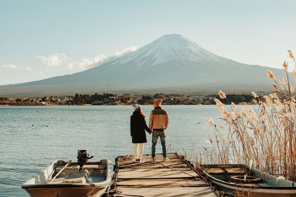 Two figures stand on a wooden dock overlooking Mount Fuji and Lake Kawaguchi in Japan with autumn reeds swaying nearby.
