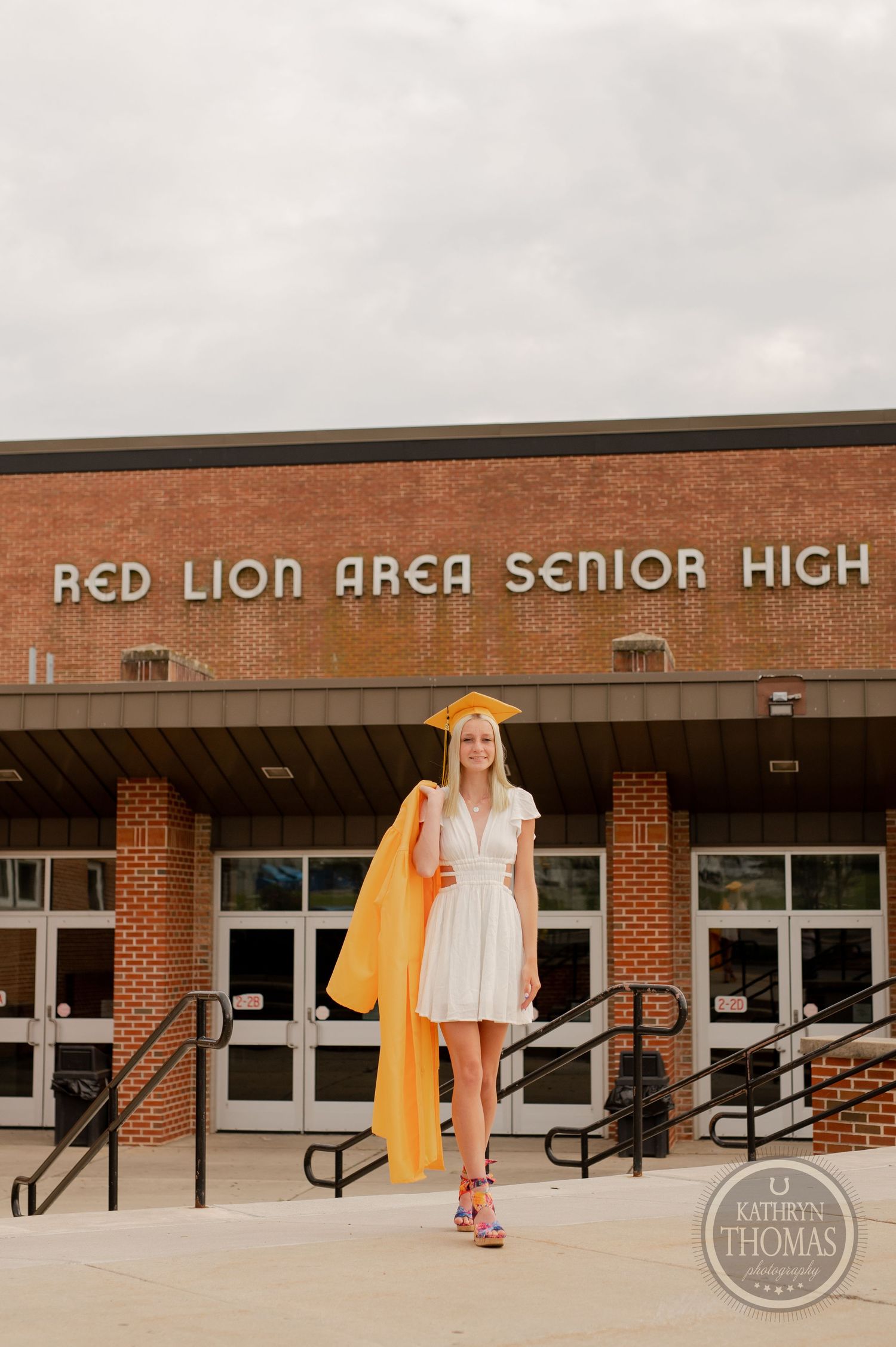 Graduate in white dress and yellow cap and gown poses in front of Red Lion Area Senior High School.