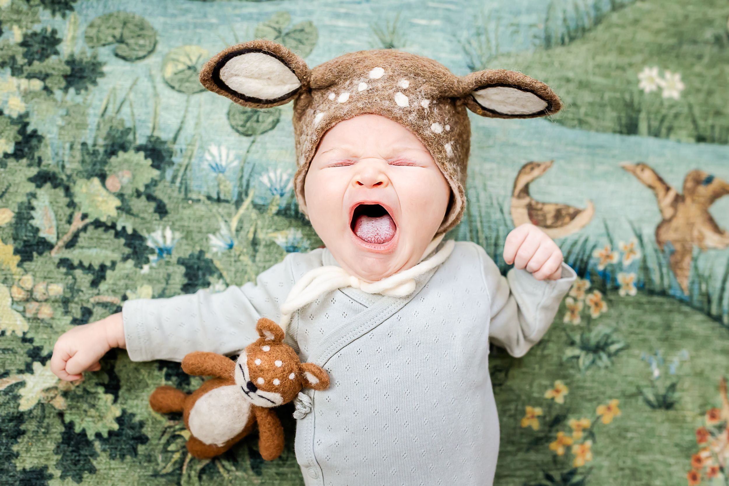 Baby wearing deer hat yawning against whimsical forest backdrop.