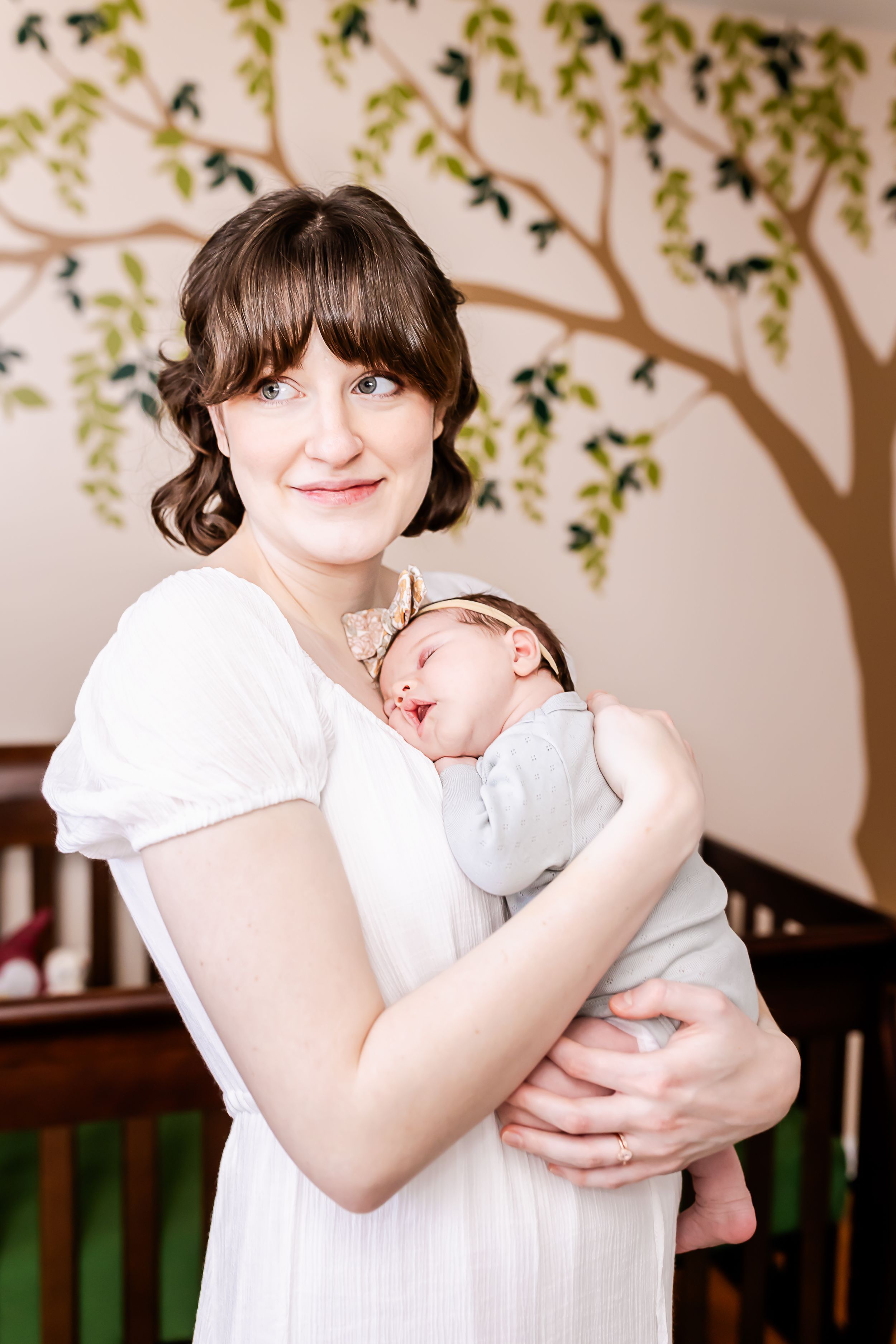 Mother holding sleeping newborn in nursery with painted tree mural.