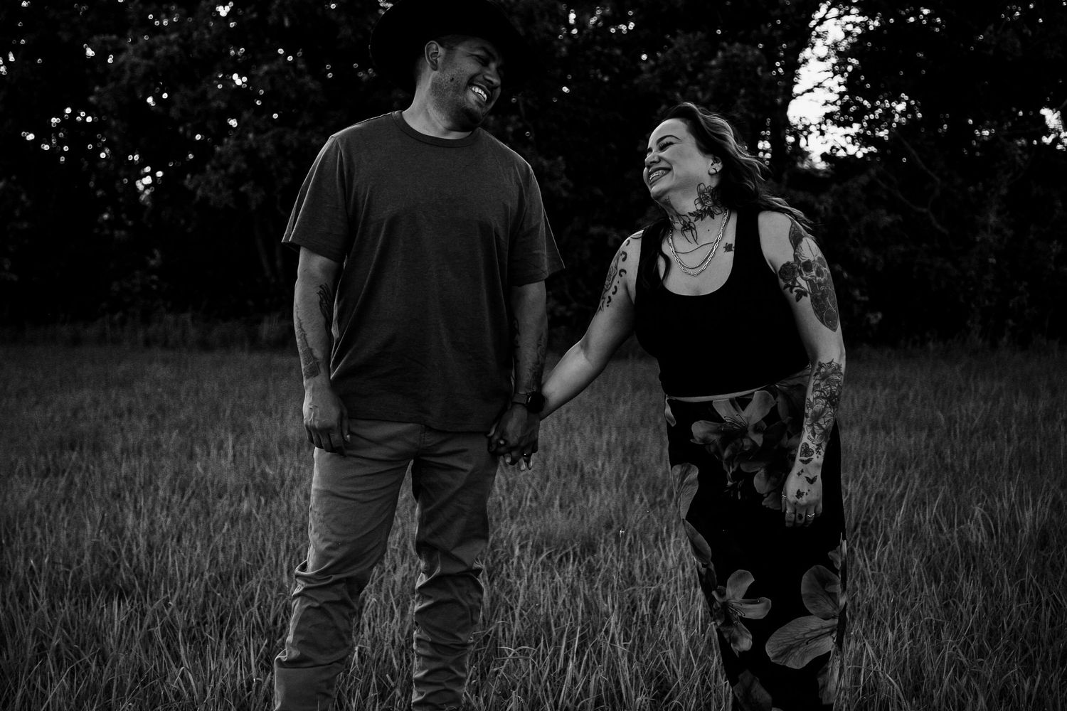 Four people hold hands while walking through tall grass in a line at sunset, with trees in the background.