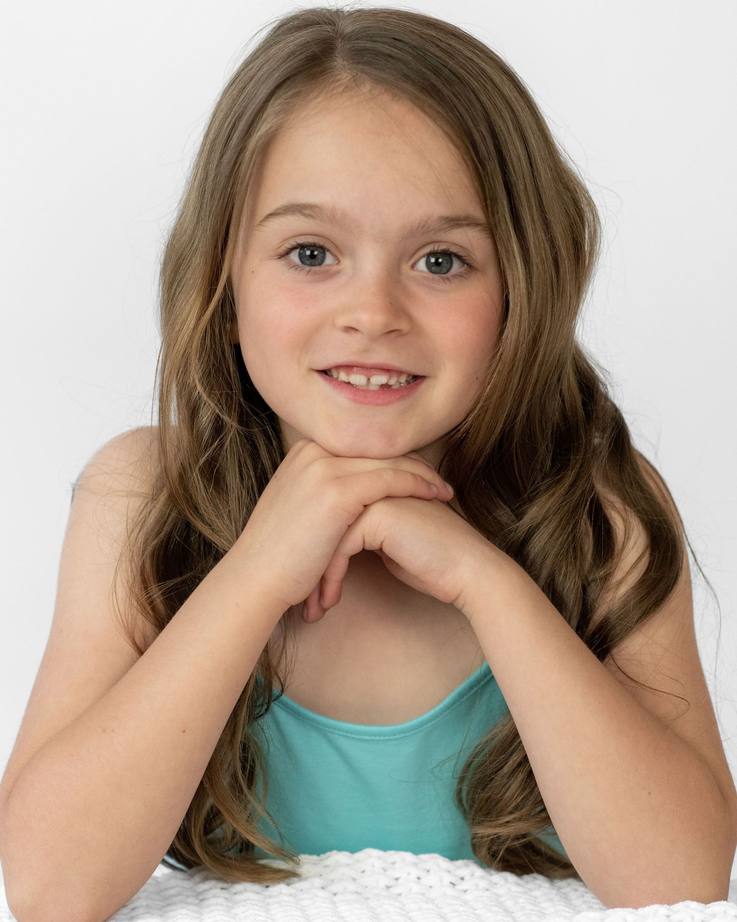 Young child in a turquoise top poses with hands under chin against white background.