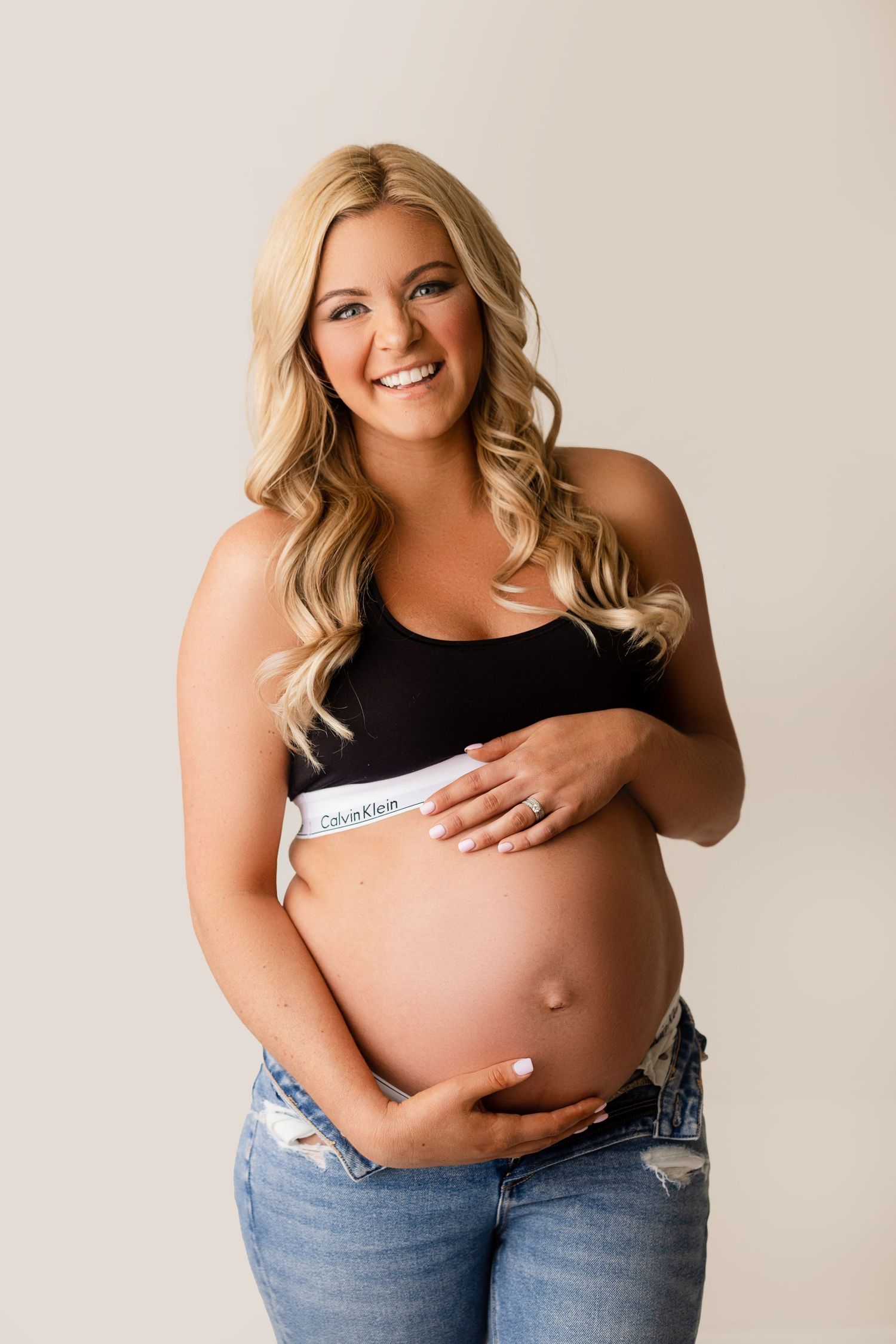 Expectant mother in black top and jeans smiles while cradling her baby bump against neutral backdrop.