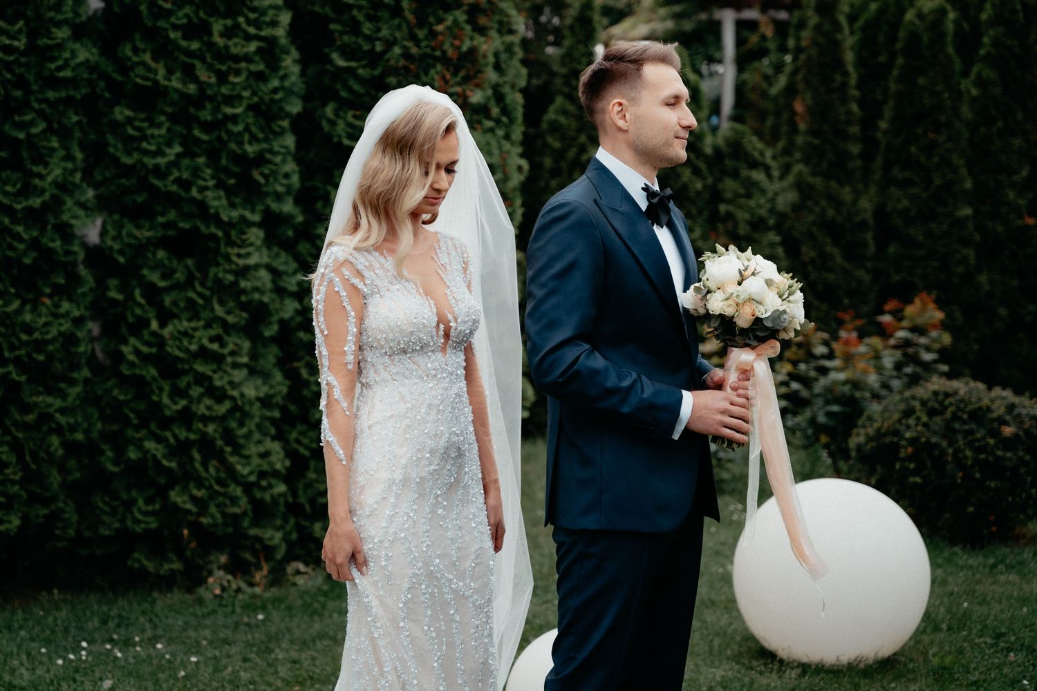 Elegant wedding couple stands in garden with groom holding white bouquet and bride wearing lace gown with balloon.