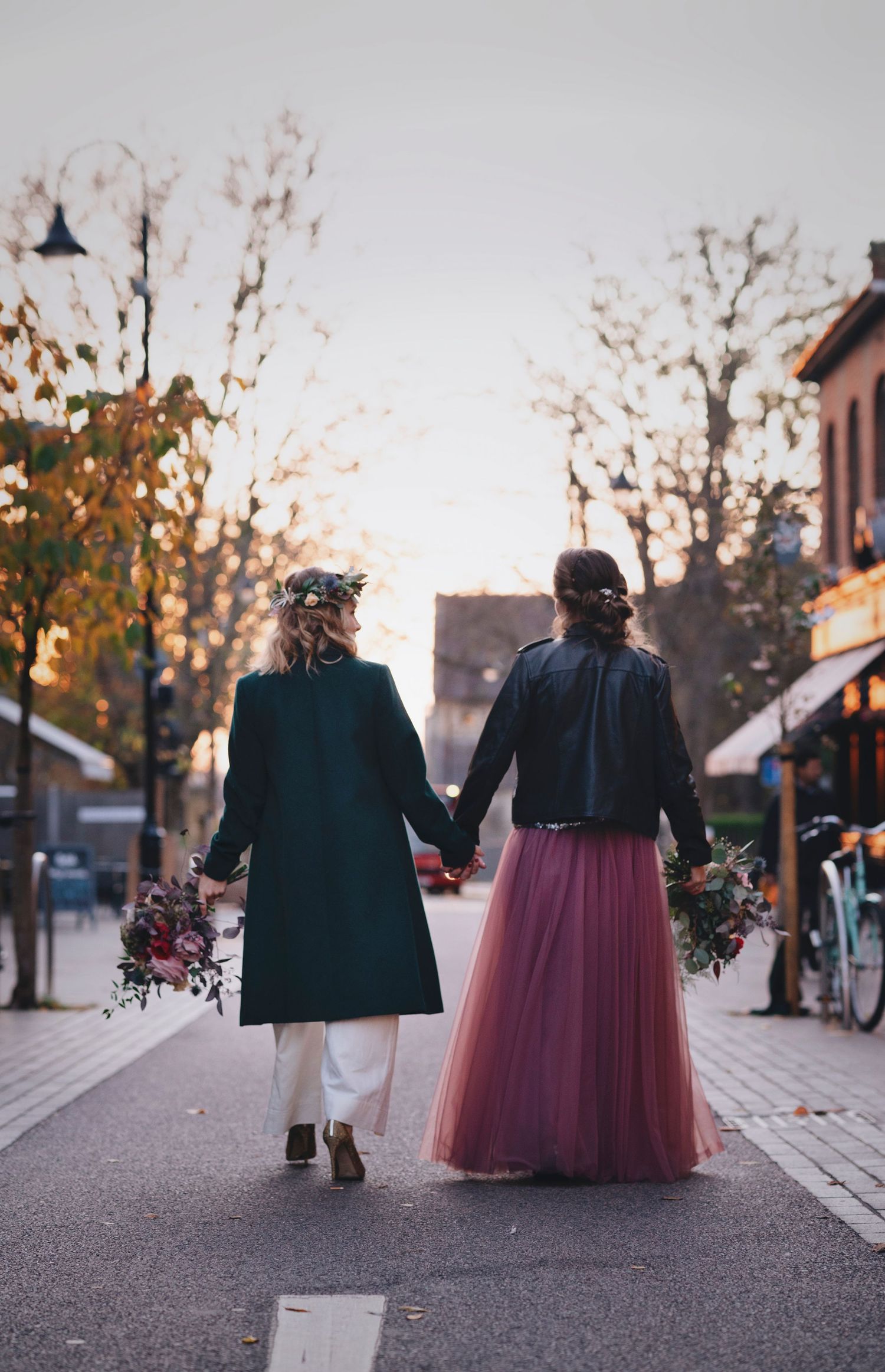 Two people in elegant attire walk hand in hand holding bouquets on an autumn street.
