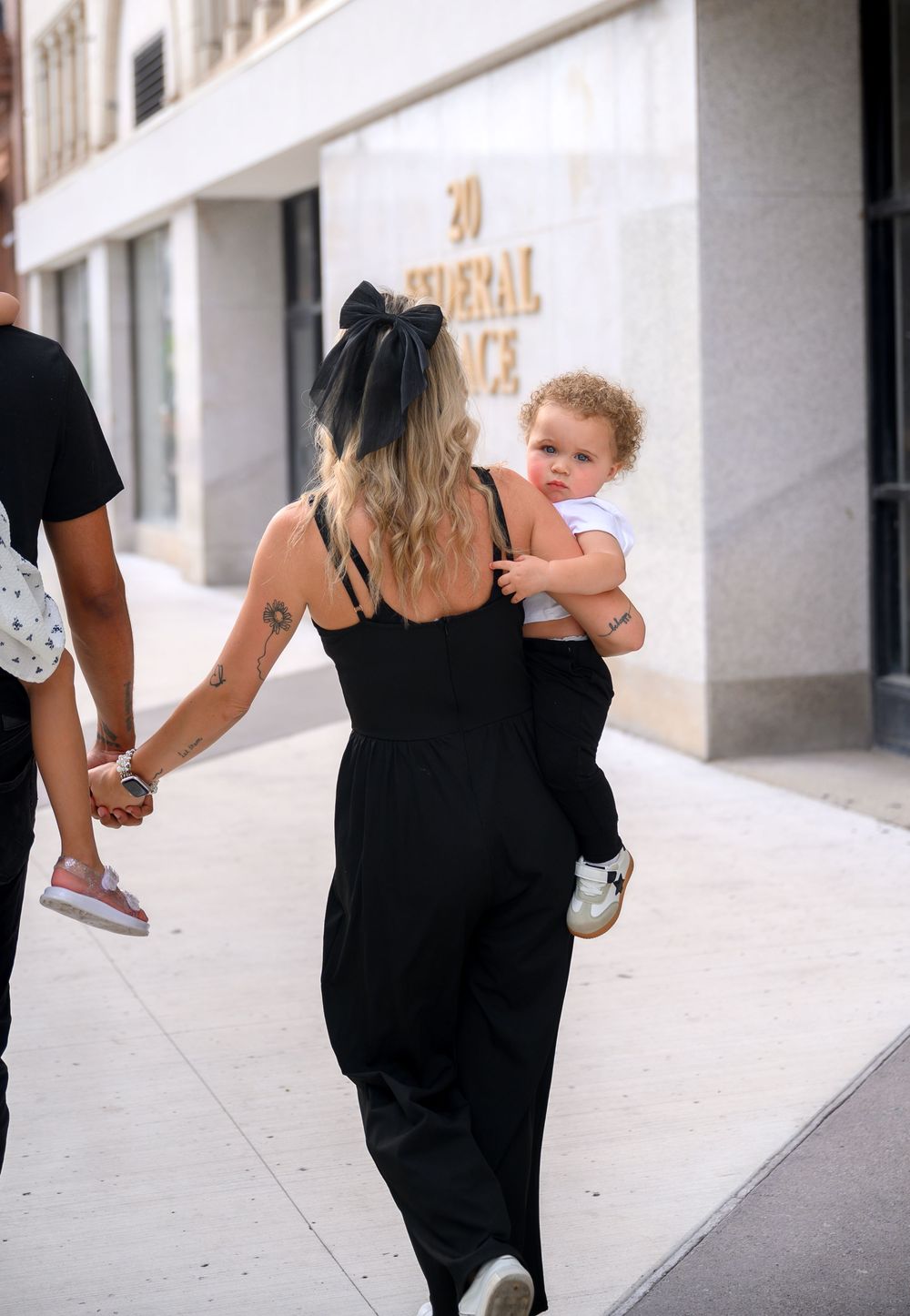A person in a black dress carries a curly-haired toddler while walking down a city sidewalk.