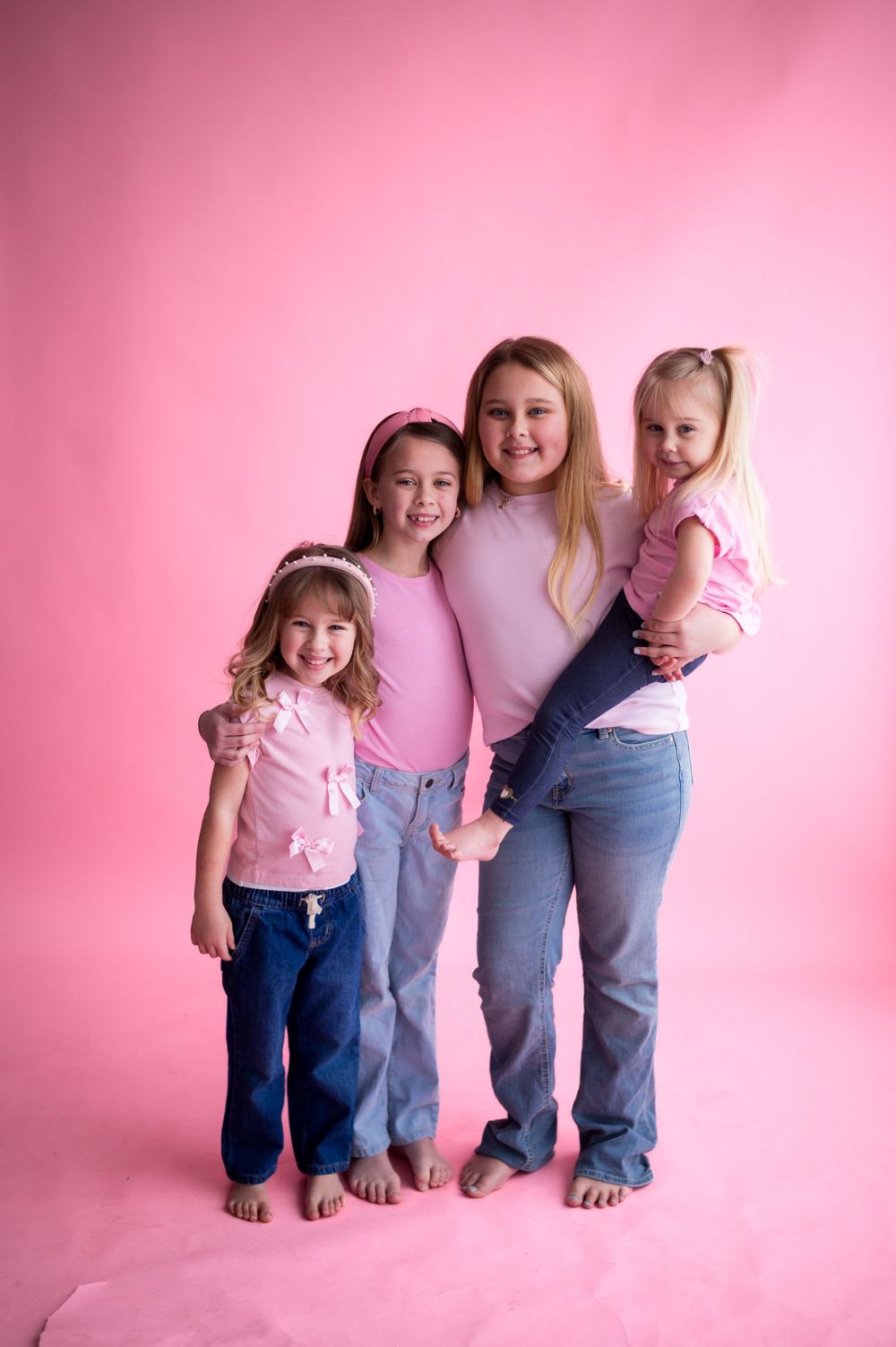 Three people wearing jeans and pink tops stand together against a pink background showing only their lower bodies.