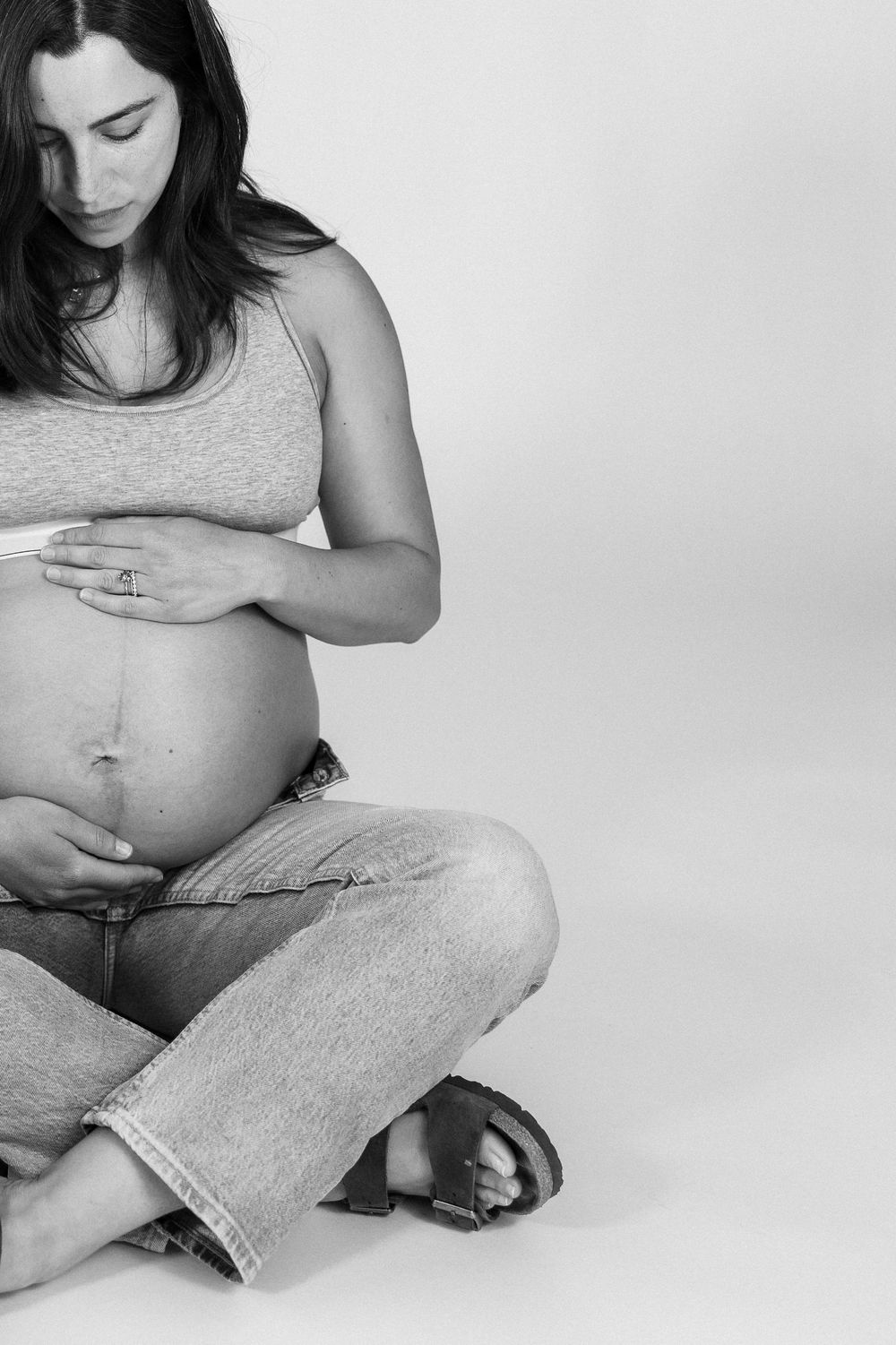 Black and white photo of a pregnant woman sitting cross-legged, cradling her bare baby bump in a tank top and jeans.