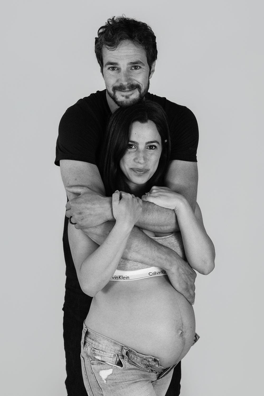 Black and white maternity photo of a smiling couple, man embracing pregnant woman from behind in studio.