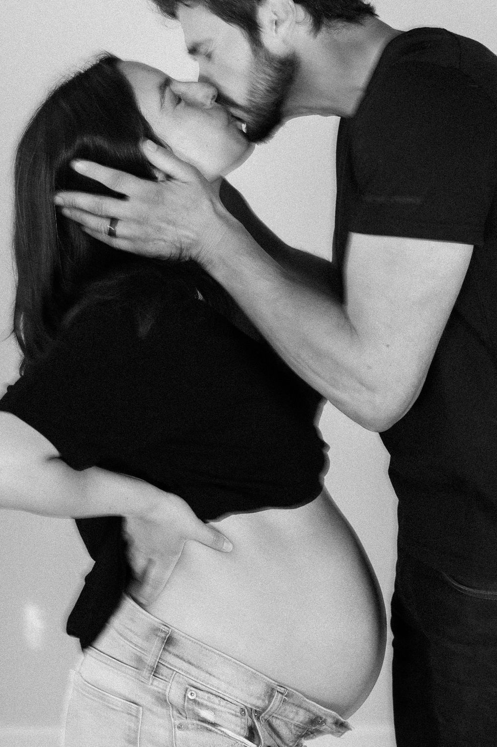 Black and white maternity photo of a pregnant couple sharing a tender kiss, woman cradling her baby bump.